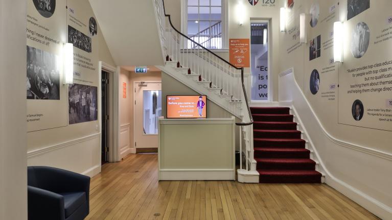 An interior hallway of Ruskin College in Oxford revealing a carpeted staircase, wooden flooring and images on the white walls