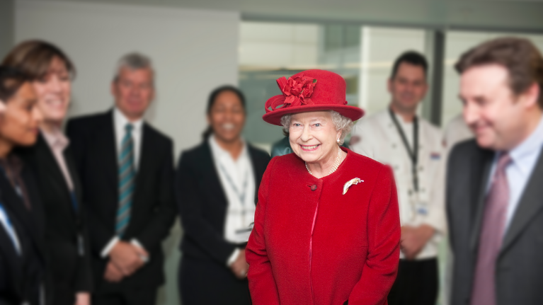 The Queen on a visit to UWL's Brentford campus. She is wearing a red hat, a red suit and a pearl necklace.