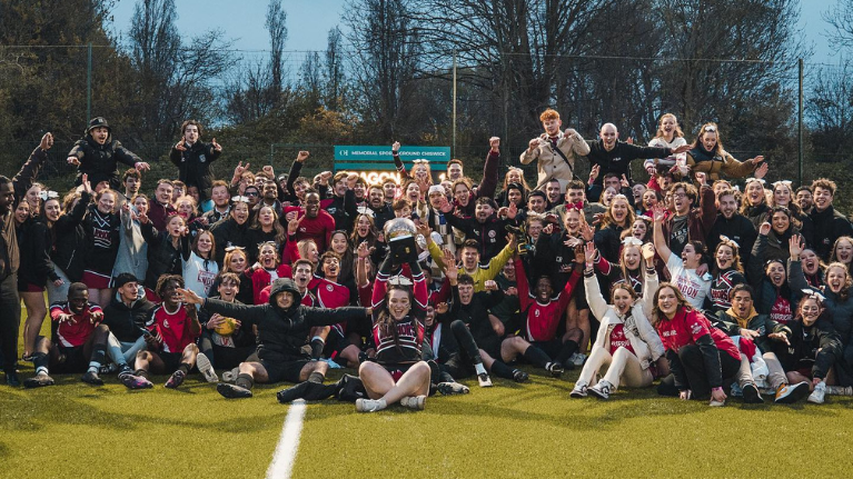 A large group of varsity sports students celebrate on a sports pitch. One student is holding a trophy above her head.