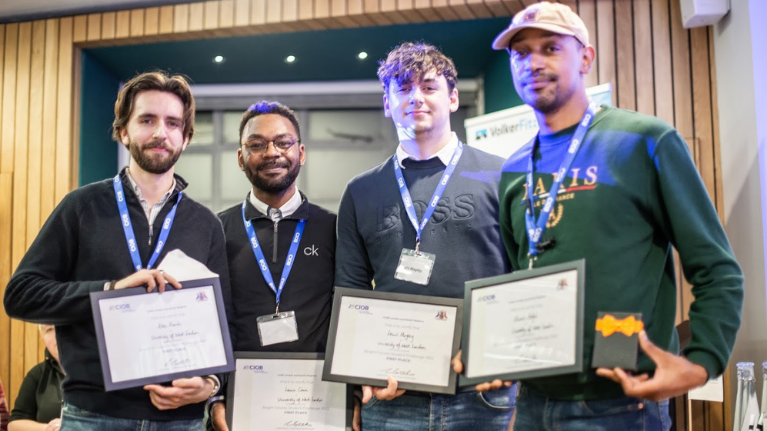 Four male students holding framed certificates. One is wearing a green jumper and the others are wearing grey jumpers.