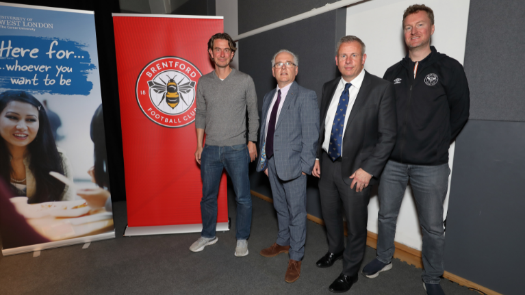 Four men are standing next to a poster of the Brentford football club logo. Two men are wearing suits and the other two are wearing jeans and a jumper. 