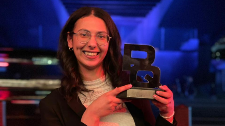 Sarah Radi is holding an award and is smiling into the camera. She is wearing a white top and black jacket, she has long brown hair and is wearing glasses.
