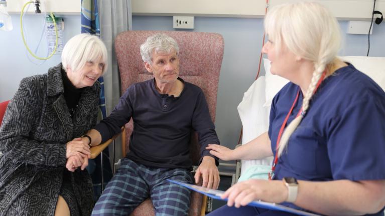 Man in hospital chair with companion talking to nurse