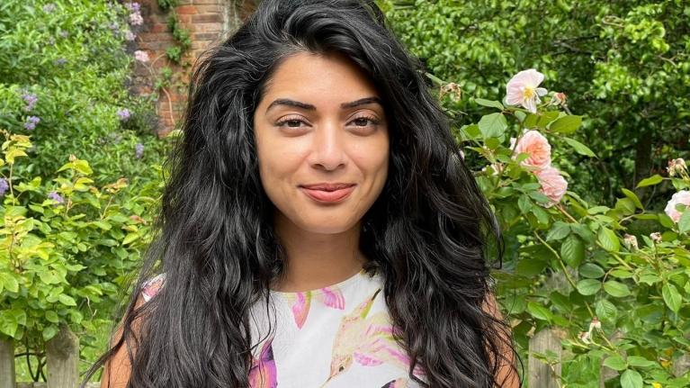 Gagan is standing infront of a rose bush and is wearing a white top with a hummingbird pattern on. She has long dark hair and brown eyes. She has a friendly smile.