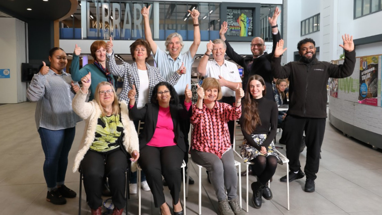 The library team are sat in the heartspace of UWL. Four are sat on chairs and 7 people are stood behind. They all have their arms lifted in triumph and are smiling.