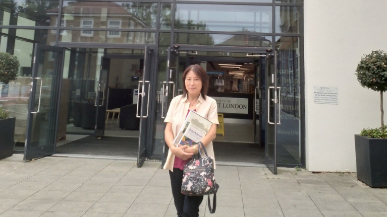 Hitomi is standing outside the park entrance to the UWL building. She is wearing a cream silk shirt over a pink t-shirt and is holding a rucksack. 