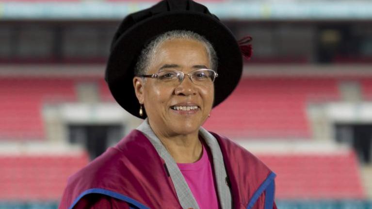 Dame Elizabeth Anionwu is wearing the UWL honorary robes and black hat. Twickenham stadium can be seen behind her. She is smiling and wearing a pink top with a grey jacket under the robes.