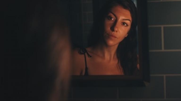Jessie is looking at her reflection in a mirror. The tiles are a sage green and her face is lit by an orange glow.