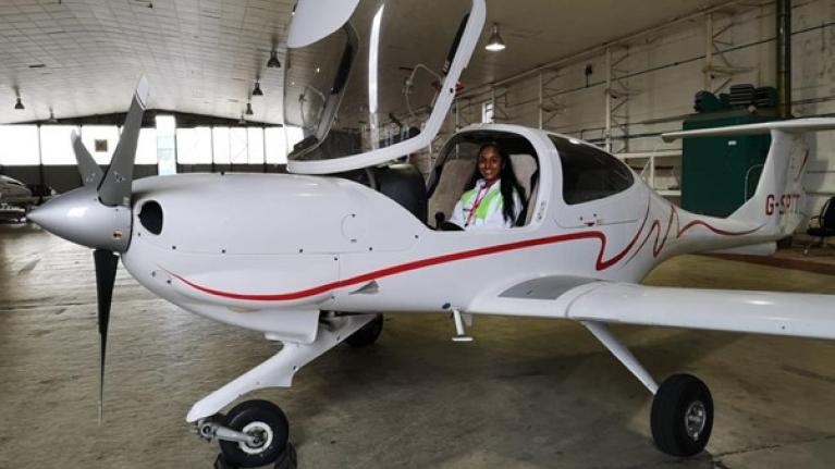 Ria Patel is smiling and sat in a small white airplane, wearing a white jacket. 
