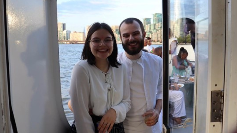 A couple are standing on a boat with a view of a city skyline behind them. They are both wearing white and both have dark hair.