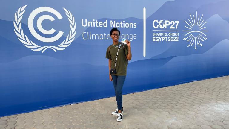 Aleigha General attends COP27, she is stood in front of a large blue screen with COP27 written on.