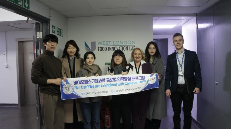 Five South Korean BioHealth Regulatory Science researchers, with Dr Amalia Tsiami and Sebastian Berg, holding a sign outside the West London Food Innovation Centre (WLFIC) stating "'We can' (We are in England with Campden BRI)".