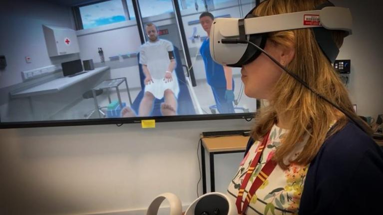 A student is wearing a VR headset with a screen behind showing two computer generated patients laying down on hospital beds.