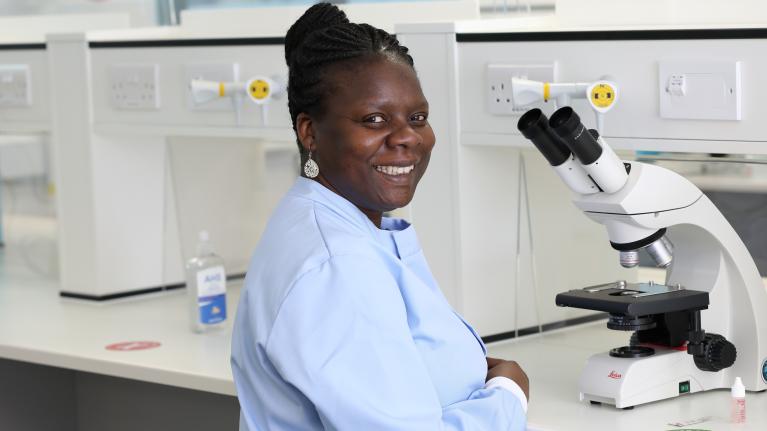 Dr Bernadine Idowu has tied up braided hair, wearing a light blue top, standing in a laboratory next to a microscope.