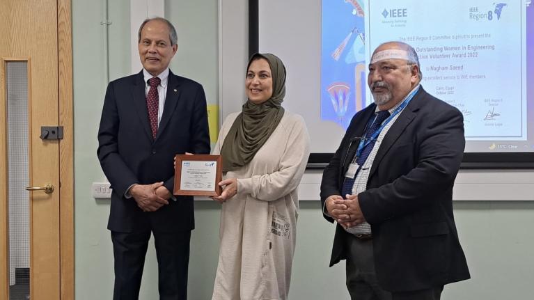 Dr Nagham Saeed holding a certificate awarding funding, standing alongside two men in suits.