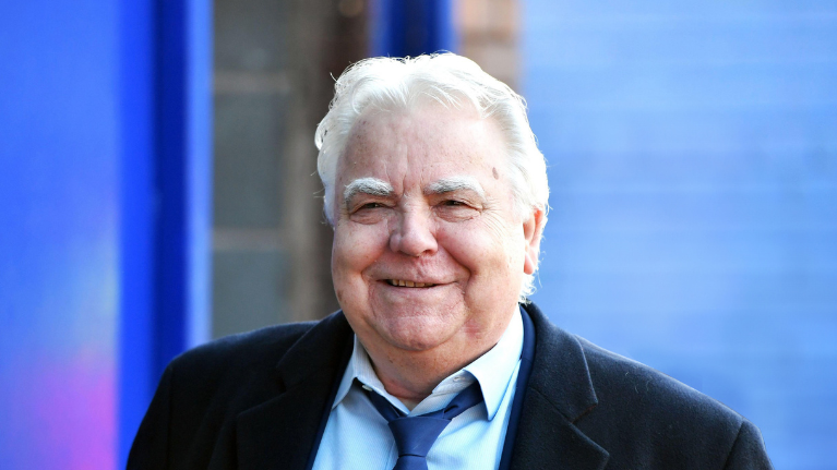 Bill Kenwright with a suit and tie on in front of a blue illuminated background.
