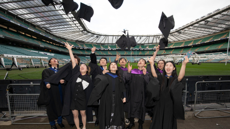 A group of graduates pitchside at Twickenham Stadium throwing their hats in the air.